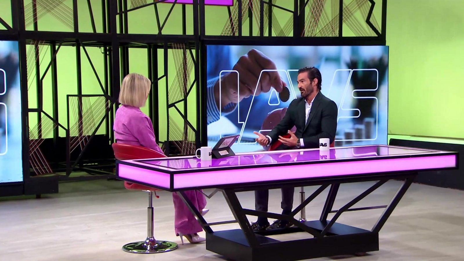 Plató de televisión con una entrevistadora con traje rosa y un entrevistado gesticulando sentados a ambos lados de una mesa iluminada con neón fucsia frente a una pantalla.