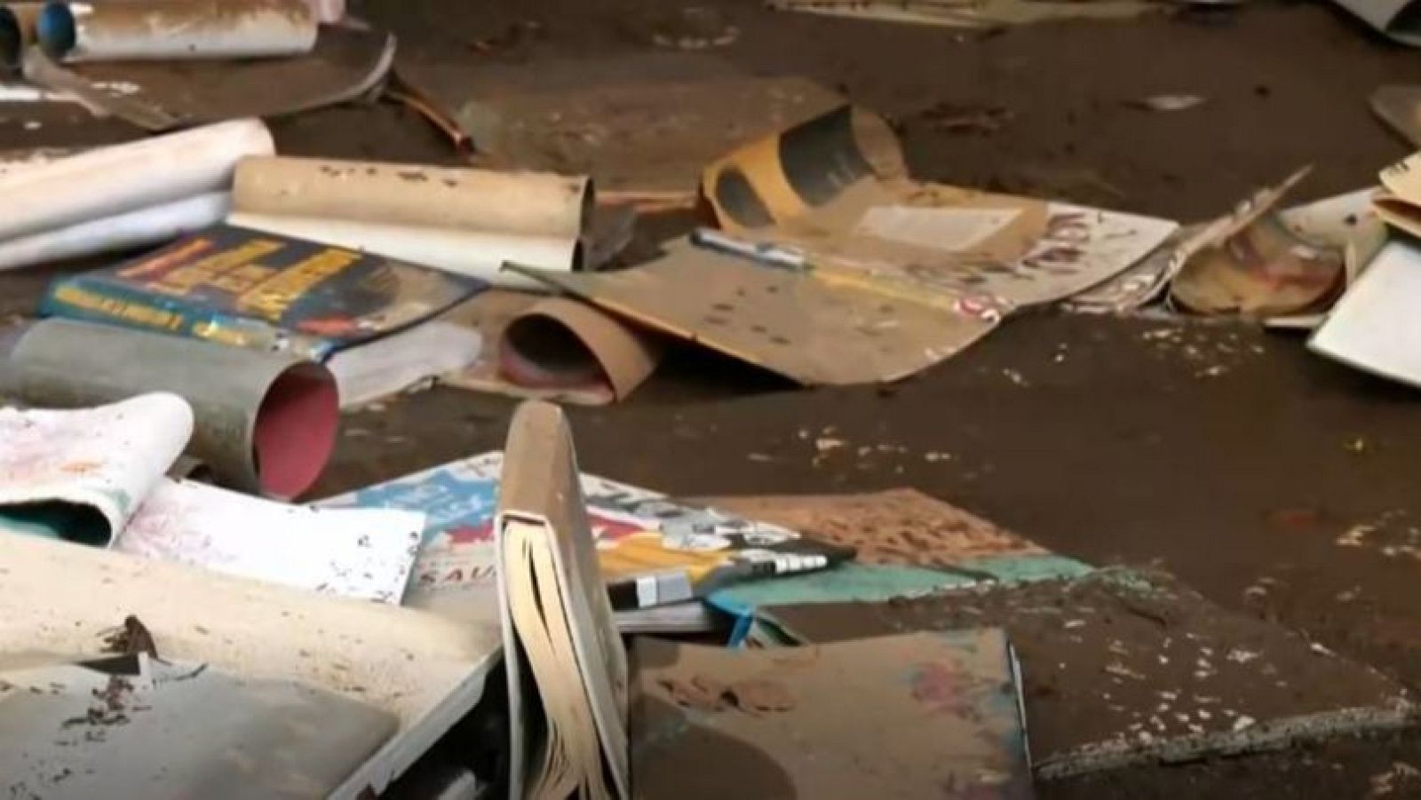 Plano cercano del suelo cubierto de lodo oscuro con libros, libretas y folios escolares arrugados y empapados de barro.