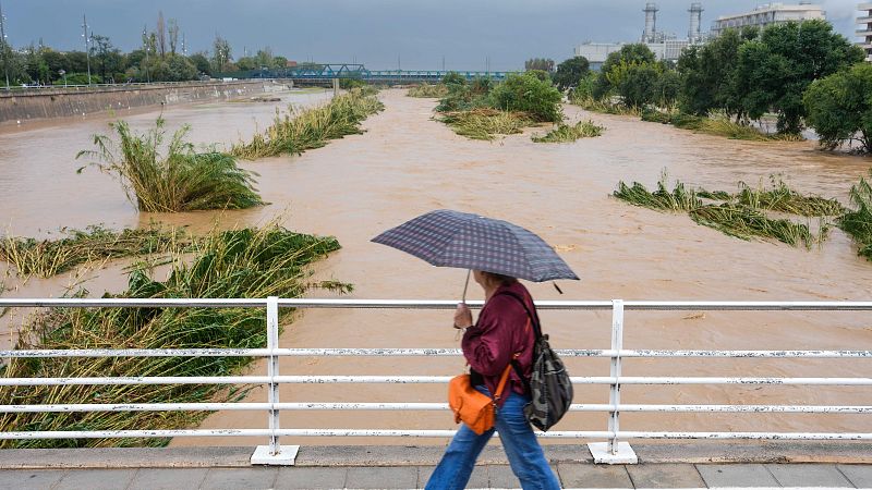 Una persona camina por un puente con paraguas junto a un río caudaloso y turbio bajo un cielo gris y lluvioso.