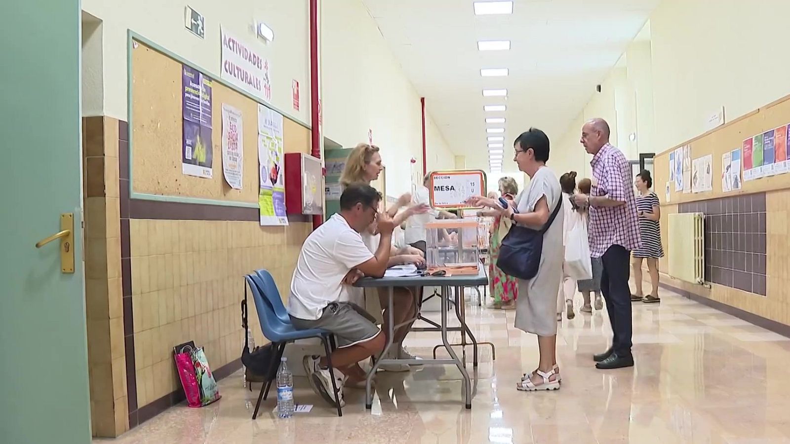 Pasillo de colegio electoral con una mesa de votación, urnas transparentes y personas esperando para votar bajo una luz fluorescente.