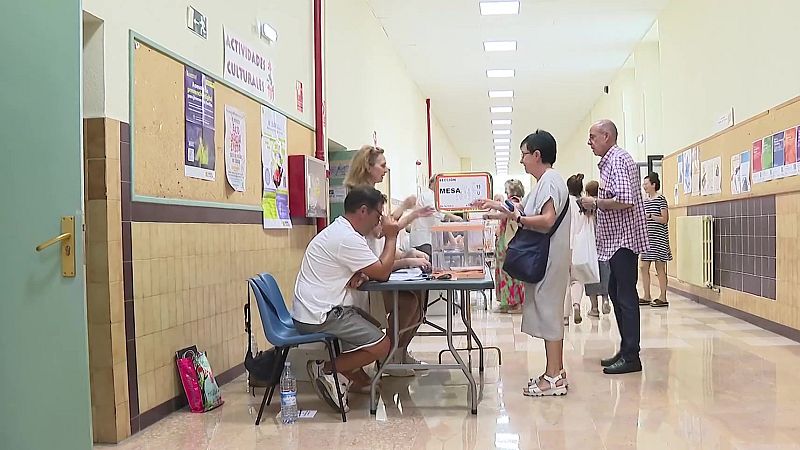 Pasillo de colegio electoral con una mesa de votación, urnas transparentes y personas esperando para votar bajo una luz fluorescente.