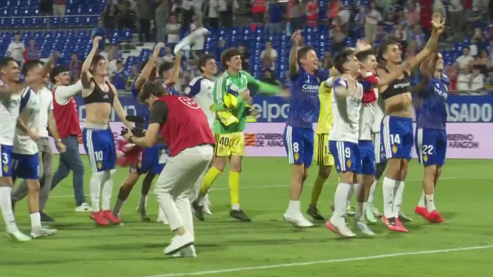 Futbolistas celebrando efusivamente sobre el césped de un estadio ante la grada, acompañados por un fotógrafo en primer plano.
