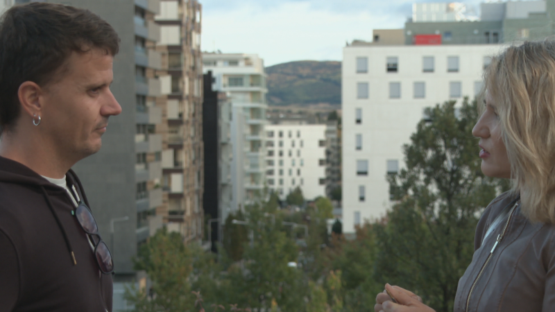 Dos personas de perfil conversan en una terraza exterior con vistas urbanas y montañosas, con un hombre escuchando y una mujer rubia gesticulando.