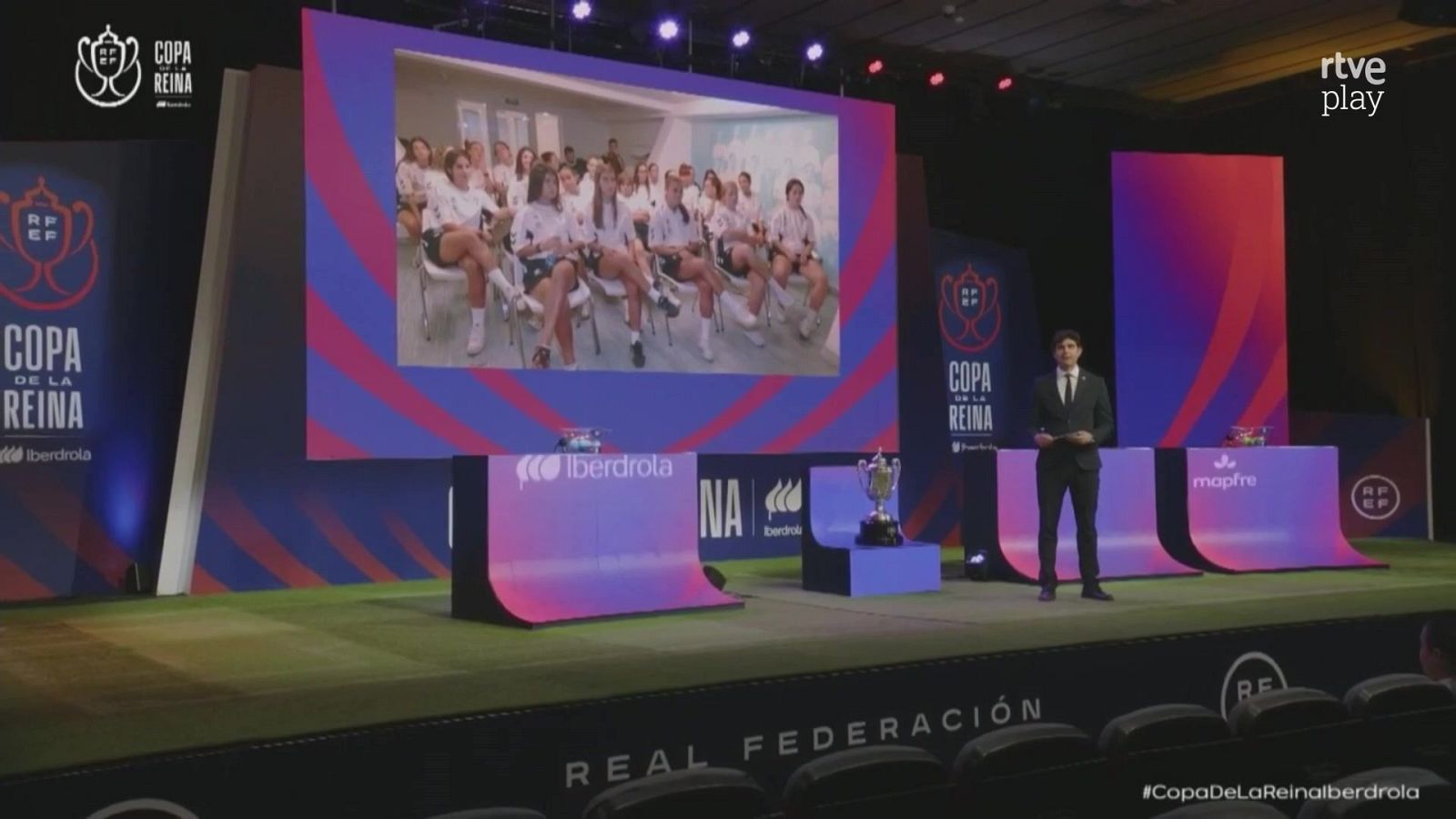 Retransmisión del sorteo de la Copa de la Reina de fútbol con el presentador en el escenario, el trofeo central y pantallas con patrocinadores.