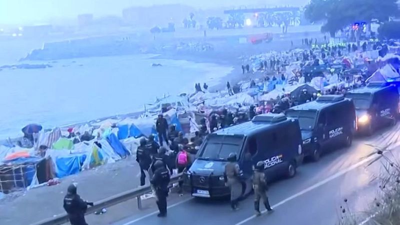 Campamento improvisado de lonas en una playa rocosa junto a una carretera custodiada por furgones y agentes antidisturbios de la policía.