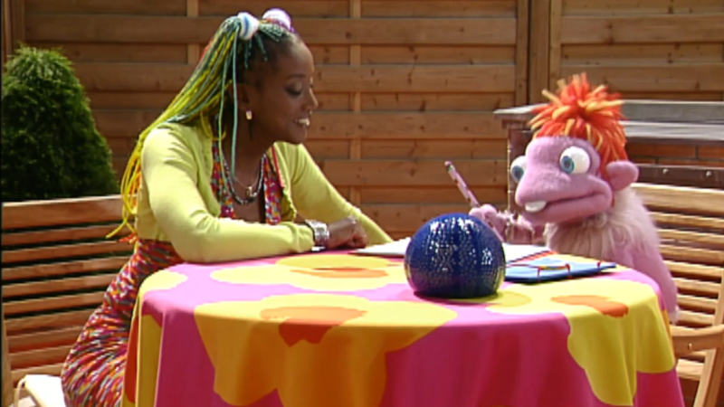 Lucrecia, con trenzas coloridas, y el títere Lucho comparten mesa en una terraza exterior mientras él dibuja en una libreta.