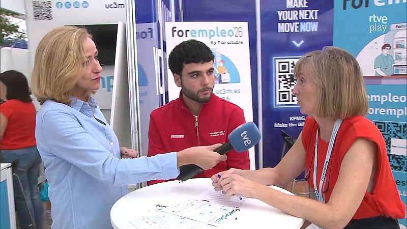 Una reportera con micrófono de Televisión Española entrevista a una mujer en un stand con folletos durante una feria de empleo, bajo la atenta mirada de un joven.