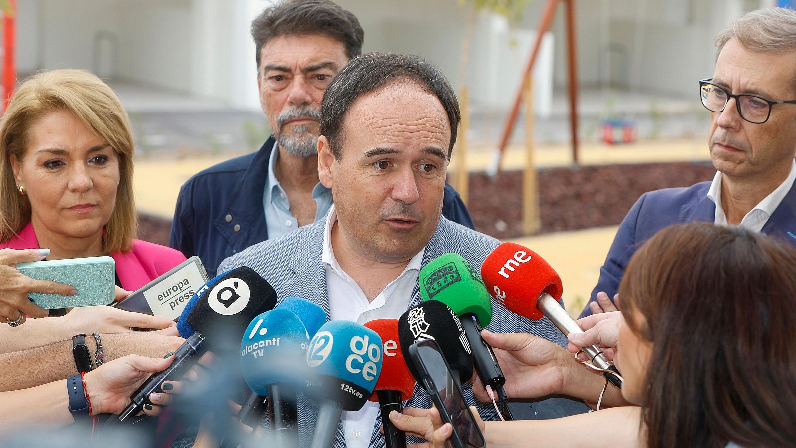 Un político con americana gris y camisa blanca habla ante una gran acumulación de micrófonos de prensa al aire libre.