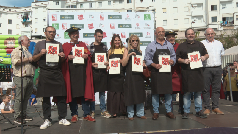 Diez personas posan en una tarima exterior frente a un photocall publicitario en un evento de txistorra, mostrando diplomas y vistiendo delantales y trajes tradicionales.