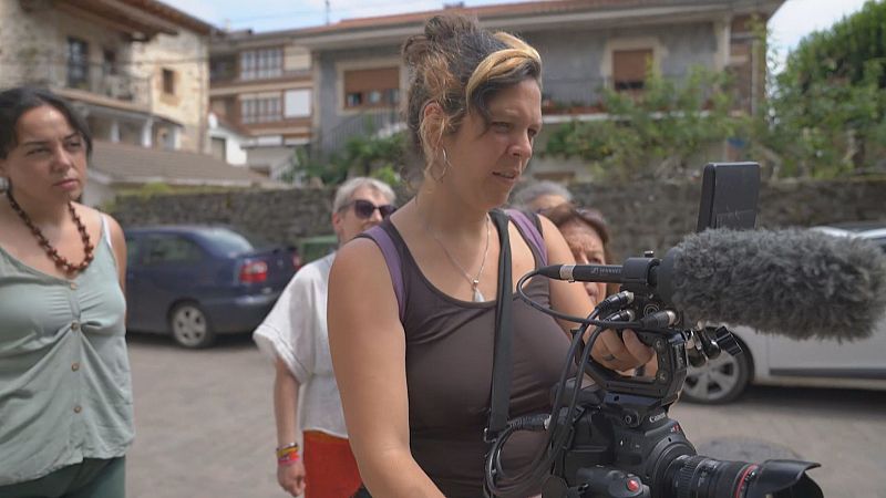 Una mujer opera una cámara de cine profesional con micrófono antiviento durante un rodaje al aire libre en un pueblo de arquitectura tradicional junto a otras participantes.