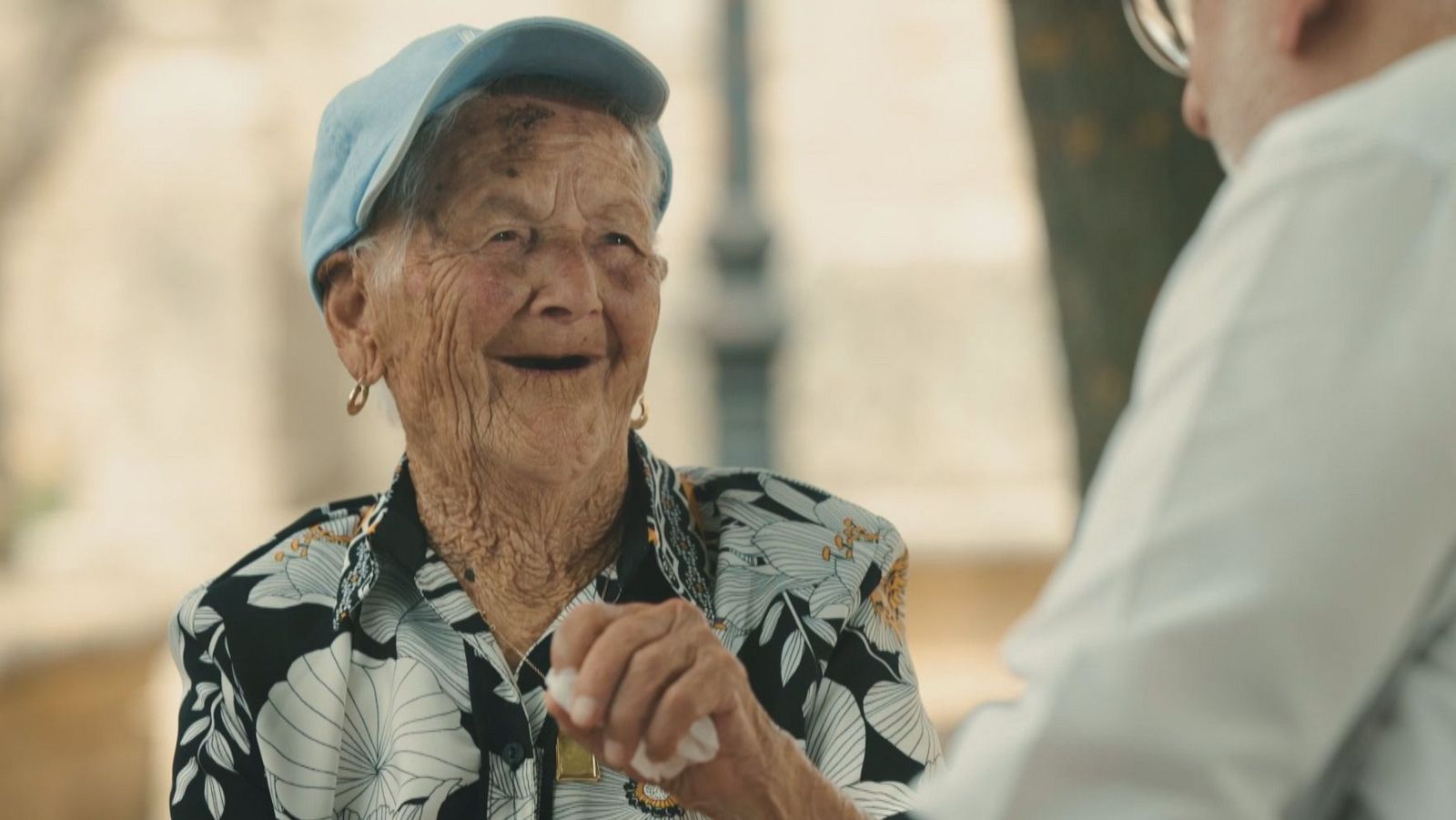Mujer de avanzada edad con sonrisa alegre, camisa estampada y gorra azul celeste conversando en exterior.