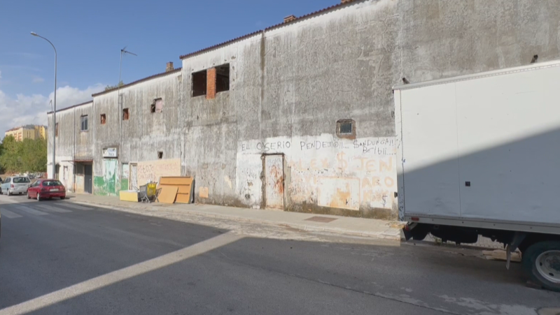 Edificio urbano abandonado de dos plantas con fachada deteriorada y grafitis, junto al que hay un camión blanco estacionado.