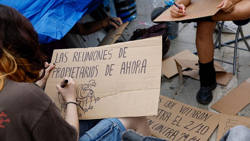Varias personas preparan pancartas de cartón en el suelo para una protesta relacionada con la vivienda, dibujando caricaturas y lemas reivindicativos.