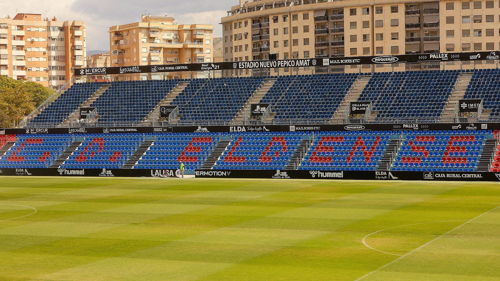 Vista del estadio de fútbol con césped en franjas verdes, gradas azules con asientos rojos que forman «C. D. ELDENSE» y un operario junto a la valla publicitaria.