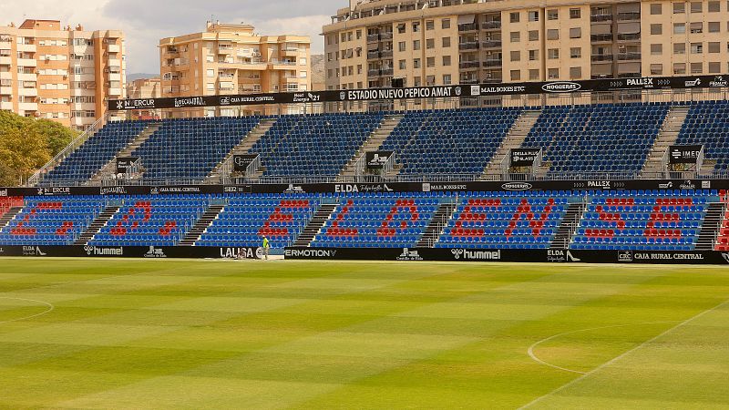 Vista del estadio de fútbol con césped en franjas verdes, gradas azules con asientos rojos que forman «C. D. ELDENSE» y un operario junto a la valla publicitaria.