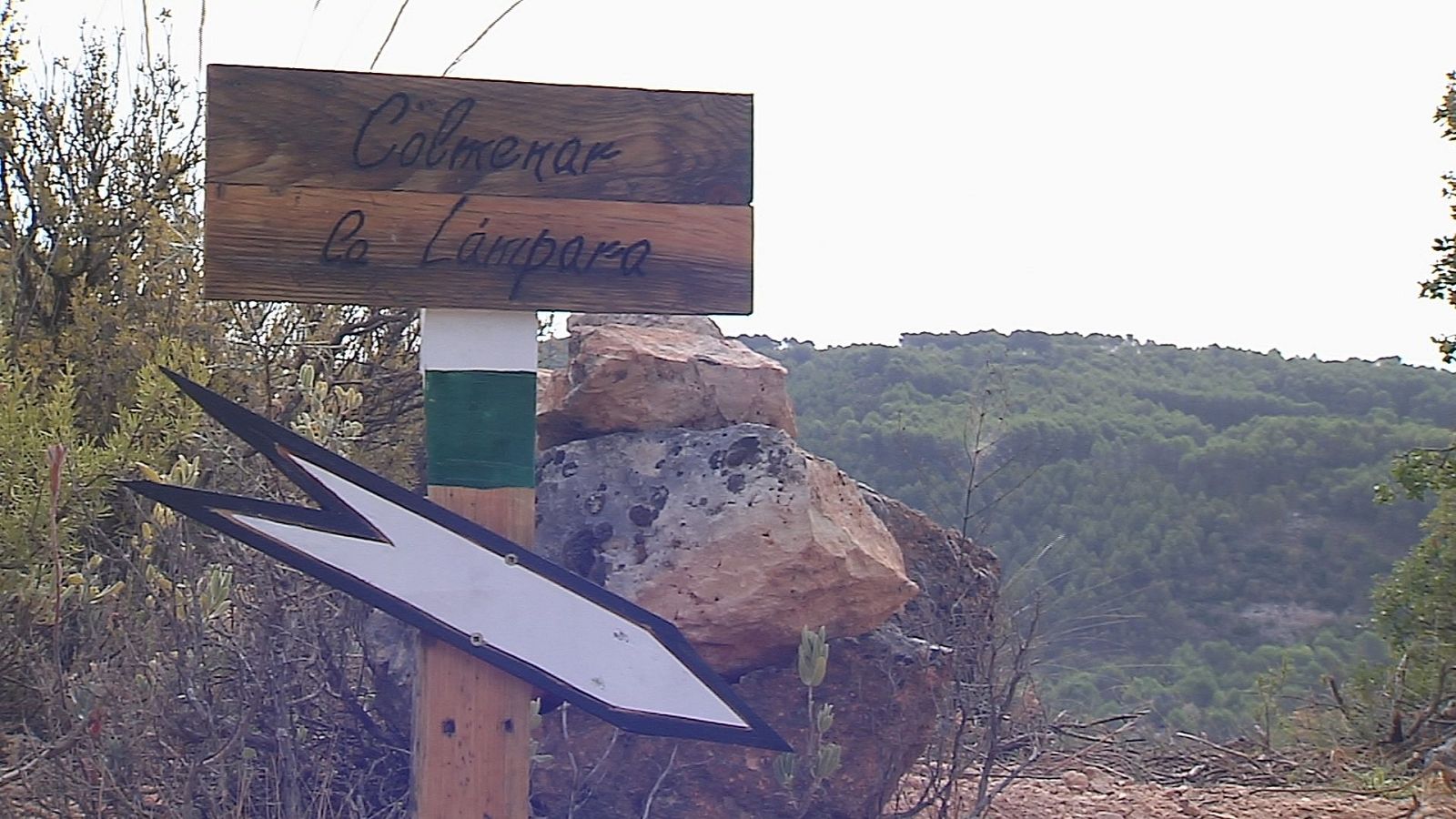 Señal de senderismo en zona montañosa con poste de madera, cartel indicador y franjas blanca y verde.