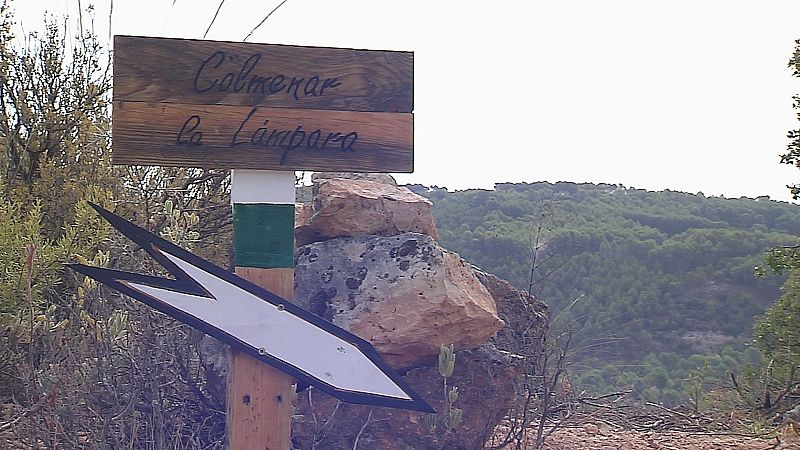 Señal de senderismo en zona montañosa con poste de madera, cartel indicador y franjas blanca y verde.