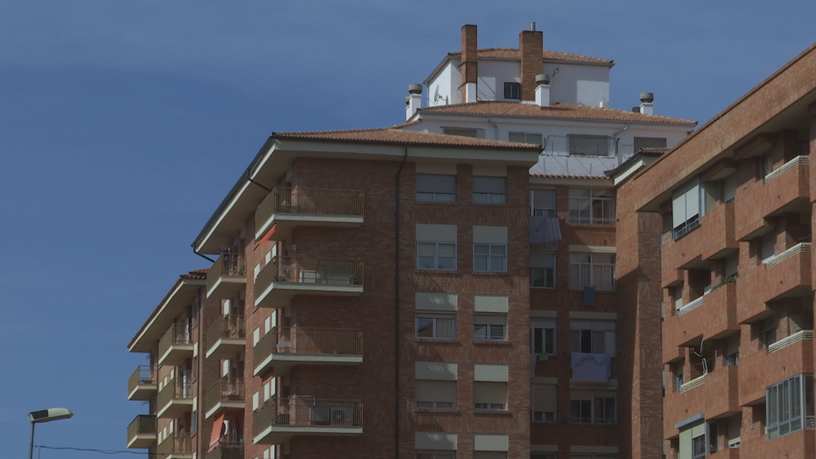 Plano contrapicado de edificios de viviendas de ladrillo rojo con balcones y persianas bajadas bajo un cielo azul.