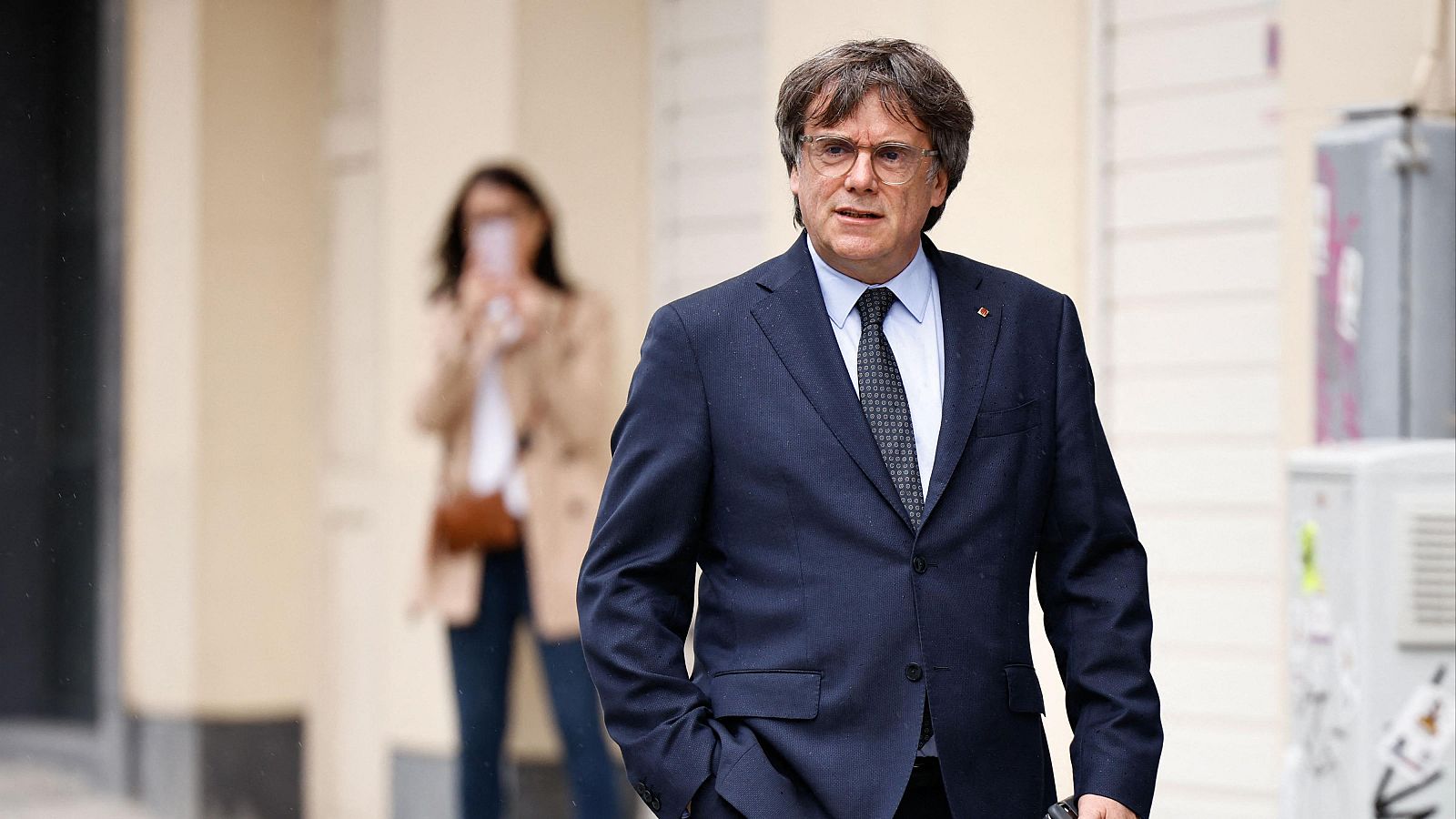 En un plano medio, el político Carles Puigdemont camina de frente por una calle con expresión seria, vistiendo un traje azul marino oscuro y corbata.
