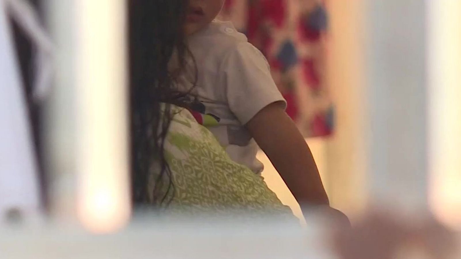 Plano cerrado de un menor de espaldas o parcialmente oculto, vistiendo camiseta blanca junto a un cojín estampado.