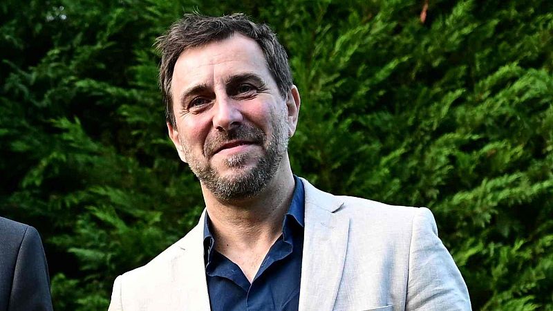 Plano medio de un hombre sonriente con barba de varios días, cabello corto y chaqueta beige, al aire libre.
