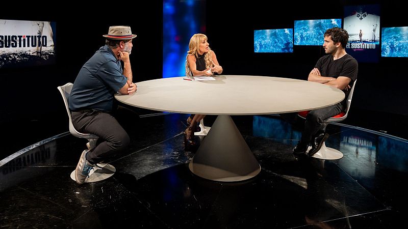 Plató de televisión con una mesa circular donde tres personas conversan sobre la película 'El sustituto' bajo una iluminación tenue y pantallas con escenas del filme.