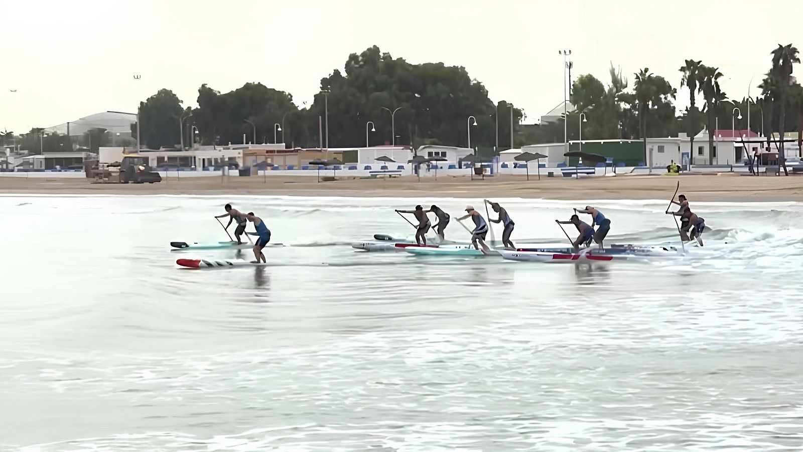Deportistas practicando paddle surf en el mar cerca de la costa, con tablas y remos, frente a una playa con paseo marítimo.