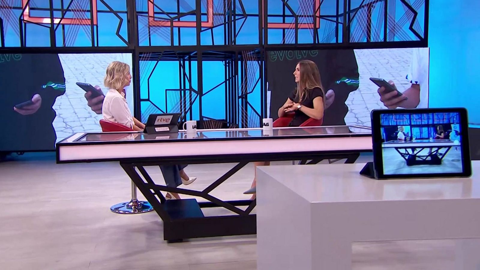 Dos mujeres conversan sentadas a una mesa moderna en un plató de televisión iluminado en tonos azules y naranjas con paneles geométricos.