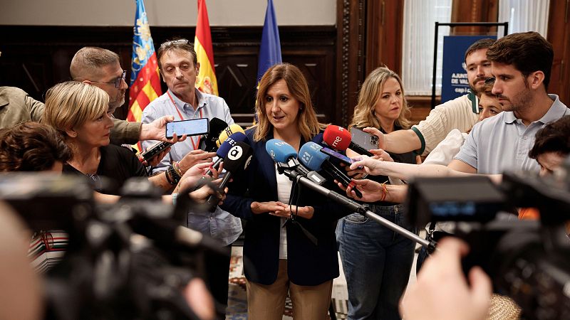 Mujer realizando declaraciones institucionales ante varios micrófonos de medios de comunicación en un espacio interior con banderas y paneles.