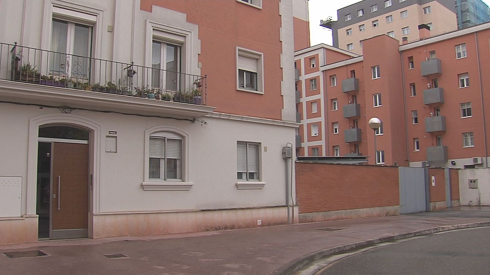 Calle residencial urbana despejada flanqueada por bloques de viviendas de color salmón con balcones y un muro de ladrillo.