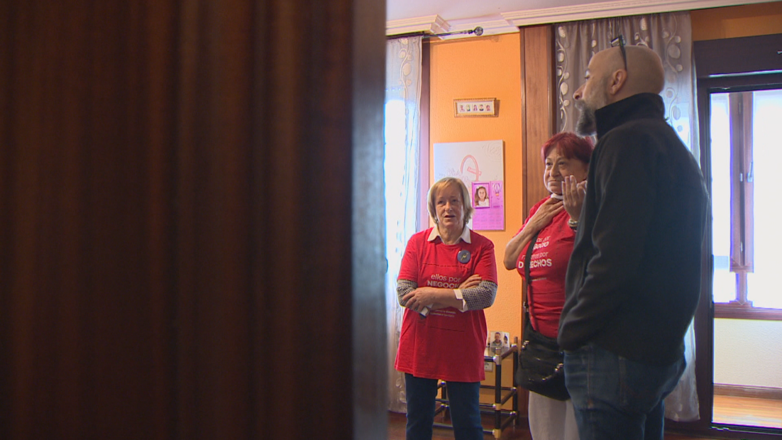 Tres personas dialogan en el salón de una casa con paredes naranjas, destacando dos mujeres con camisetas rojas y un hombre con chaqueta oscura.