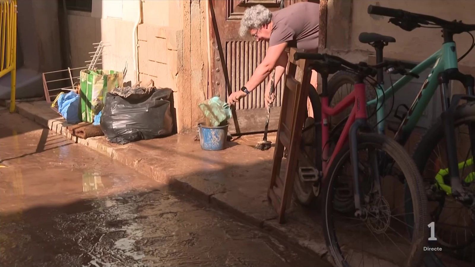 Una mujer con gafas retira agua y barro de la acera con una pala y un cubo azul tras una inundación en la calle, junto a bolsas de residuos y dos bicicletas.
