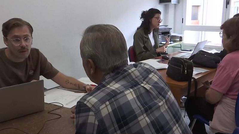 Interior de una oficina luminosa con dos puestos de atención al ciudadano donde personal administrativo atiende a usuarios frente a ordenadores y documentos.