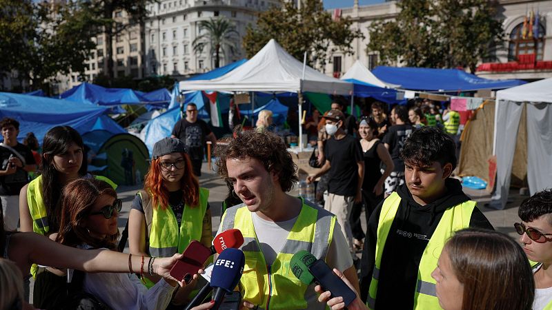 Joven con chaleco reflectante habla ante micrófonos de prensa en una protesta en la plaza con carpas y una bandera de Palestina.