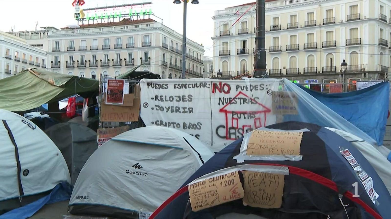 Protesta con tiendas de campaña y pancartas contra la especulación inmobiliaria en una plaza urbana frente a edificios históricos.