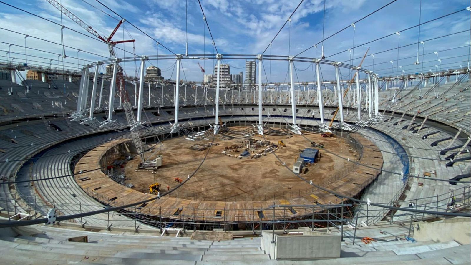 Estadio de fútbol en obras con una gran estructura circular de cubierta, terreno de tierra y grúas de construcción.