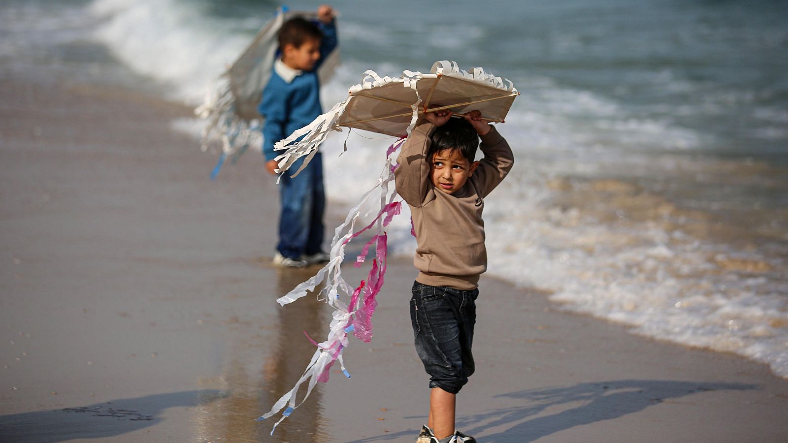 Dos niños caminan por la orilla de una playa sosteniendo cometas artesanales de plástico y varas con largas colas de colores.