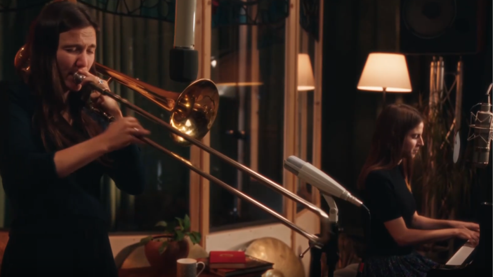 Dos músicas actúan en un estudio cálido: una mujer toca el trombón y otra interpreta el piano.