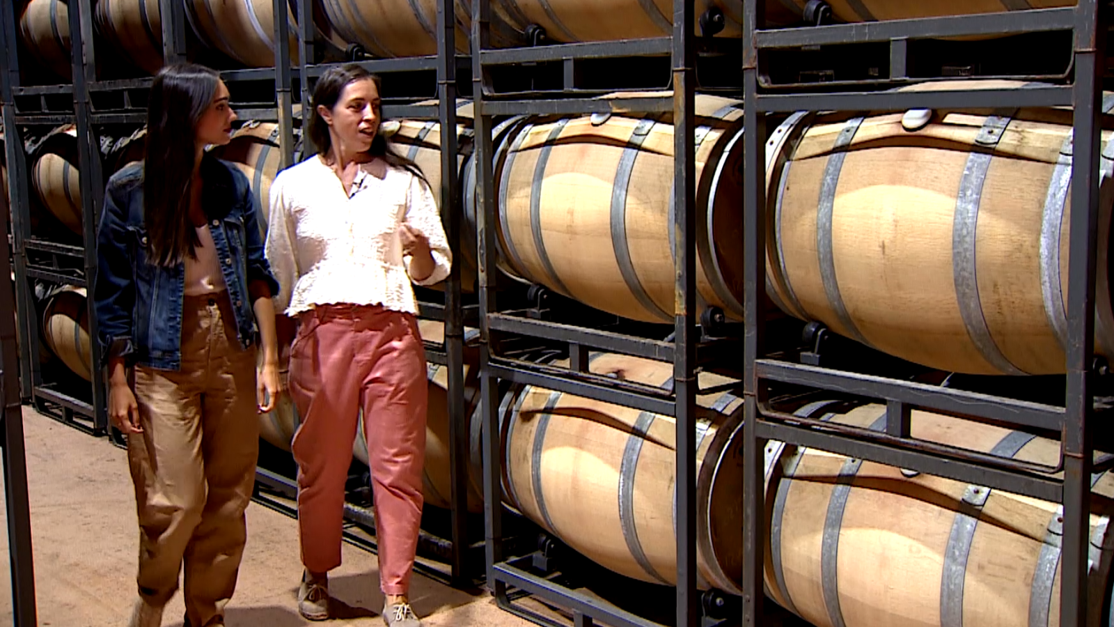Dos mujeres conversan recorriendo un pasillo de bodega iluminado cálidamente y flanqueado por estanterías metálicas con barricas de roble.