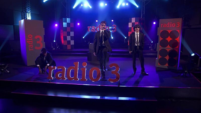 Tres m�sicos con traje oscuro y gafas de sol act�an en un escenario iluminado de azul y violeta junto al logotipo rojo del programa.