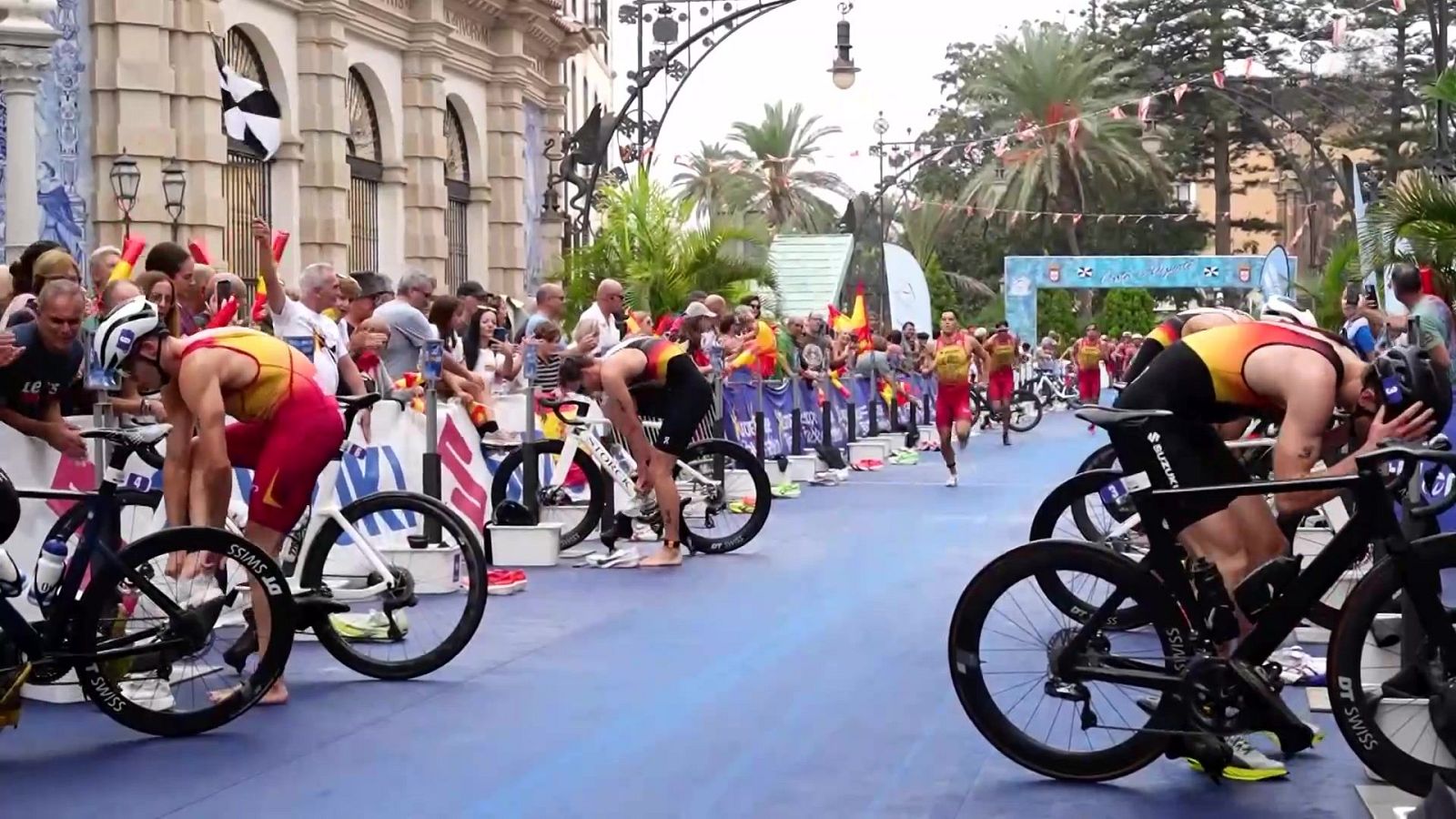 Triatletas preparándose junto a sus bicicletas en la zona de transición de una competición urbana sobre una alfombra azul.