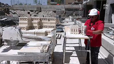 Mujer con EPI y casco blanco manipula ladrillos en el exterior de una obra con cortadora y pal� de materiales.