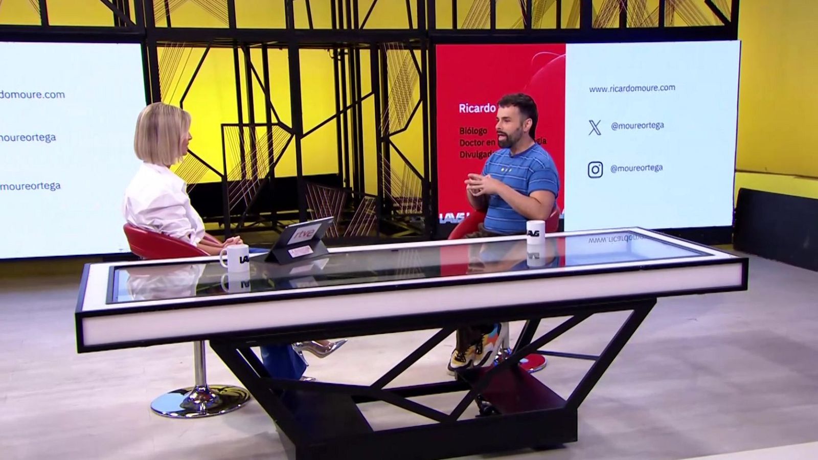 Dos personas conversan sentadas en sillas rojas frente a una mesa de cristal en un plató con fondo amarillo y pantallas informativas.