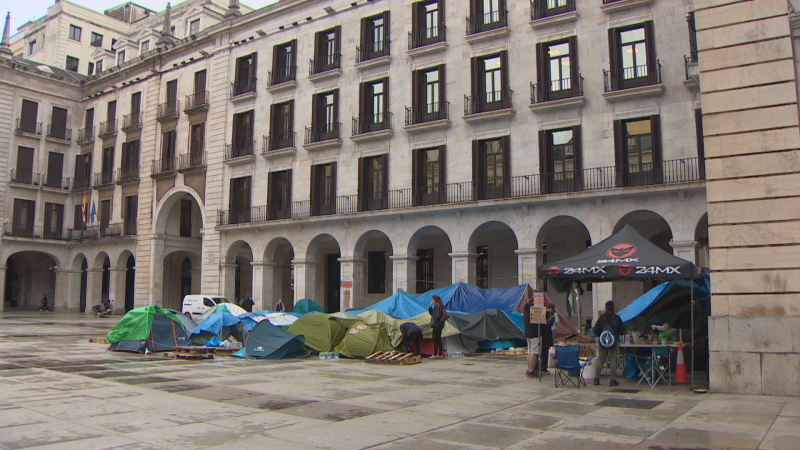 Plano general de la Plaza Porticada en Santander con una acampada de protesta formada por tiendas de campaña, lonas y una carpa negra.