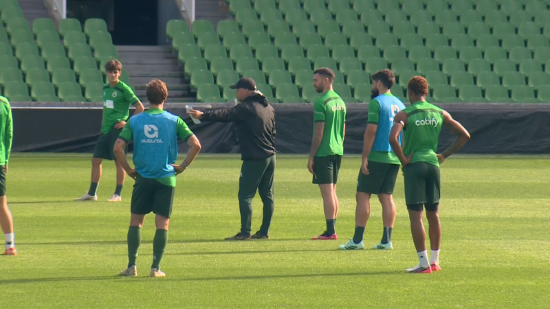 Entrenador con chaqueta negra y gorra imparte instrucciones tácticas a futbolistas en el césped de un estadio con gradas verdes vacías.