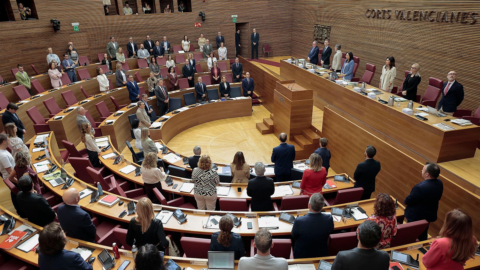 Vista en plano picado del hemiciclo de Les Corts Valencianes con los diputados de pie guardando un minuto de silencio junto a sus pupitres de madera.
