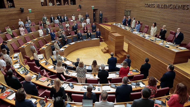 Vista en plano picado del hemiciclo de Les Corts Valencianes con los diputados de pie guardando un minuto de silencio junto a sus pupitres de madera.
