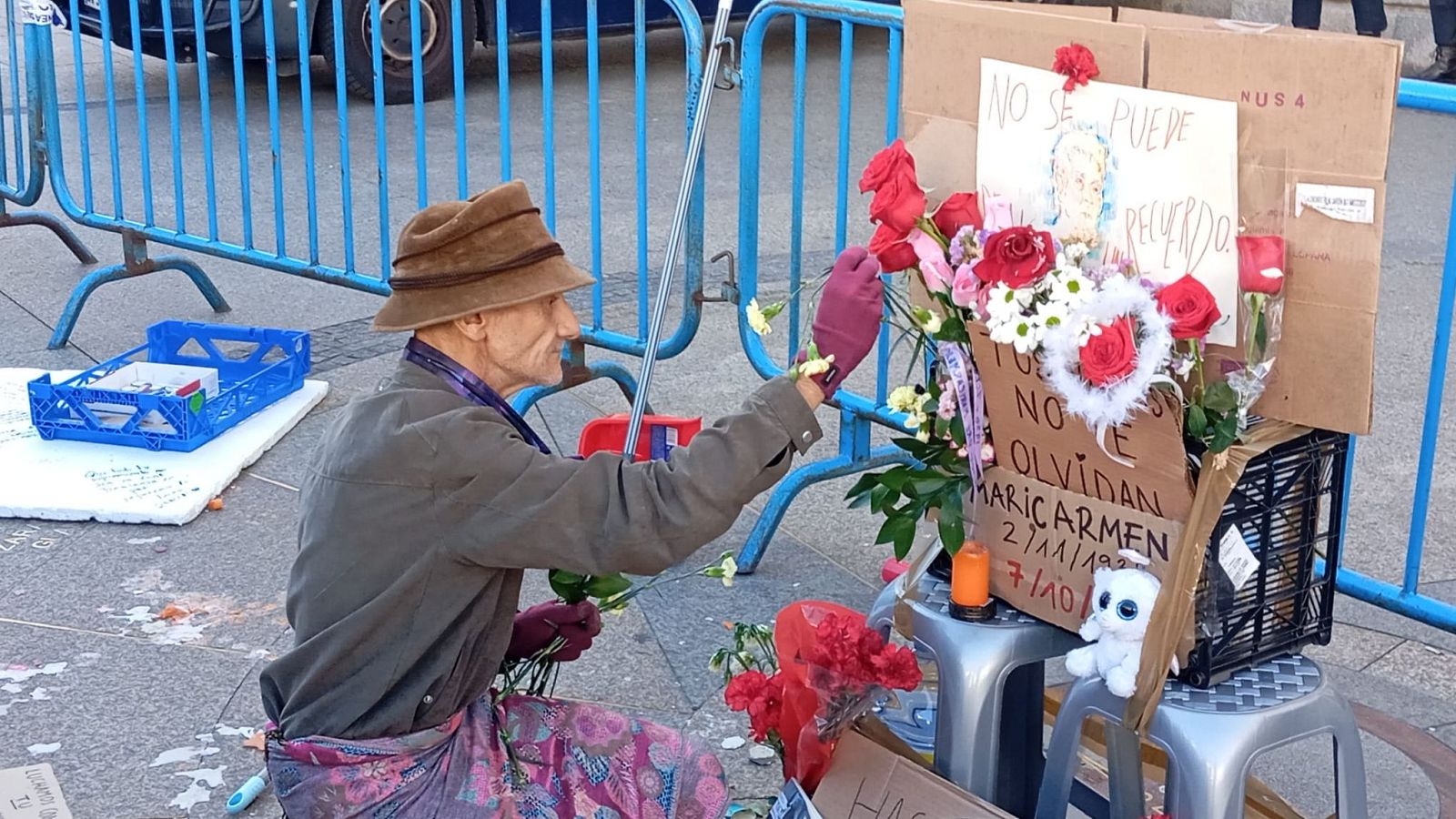 Un hombre rinde homenaje colocando una flor en un memorial improvisado con flores, velas y carteles en una plaza urbana.