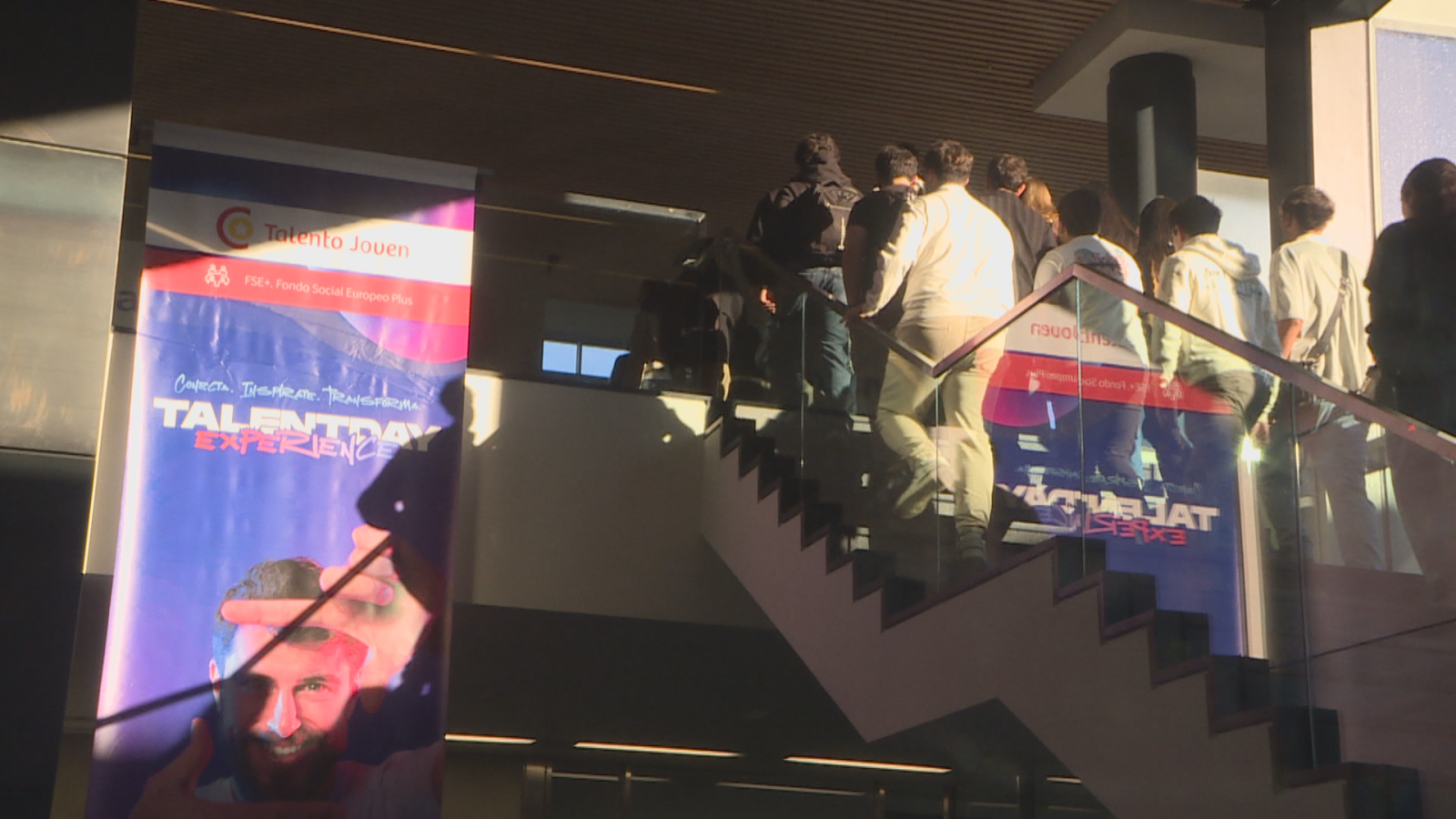 Un grupo de jóvenes sube por una escalera de diseño contemporáneo junto a un cartel publicitario de un evento de empleo.