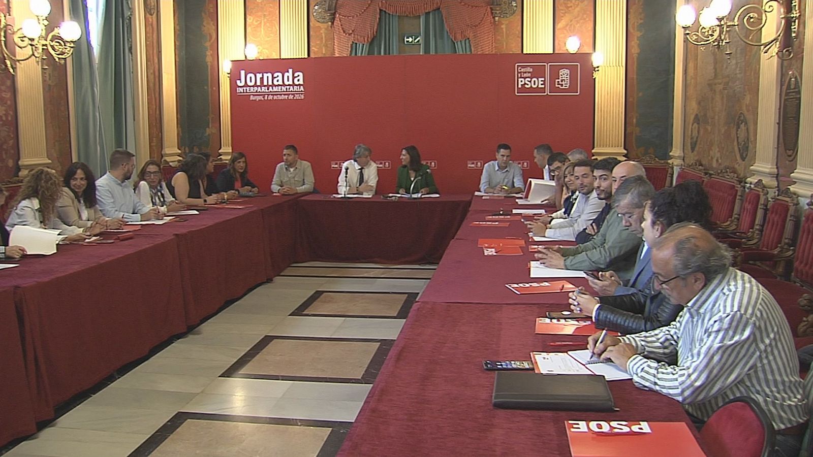 Reunión política institucional con una mesa en forma de U cubierta de mantelería granate y asistentes sentados alrededor con documentos.